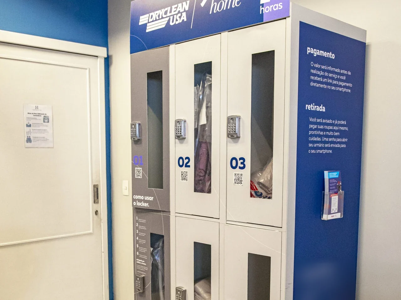 Torre de lockers DryClean USA instalada na área comum de um prédio, com portas numeradas, fechadura por teclado, QR code em cada porta e painel explicando pagamento e retirada